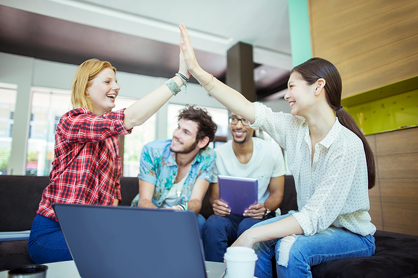 Due ragazze sedute in una zona relax dell'ufficio che si danno il cinque mentre due colleghi maschi seduti dietro di loro guardano.